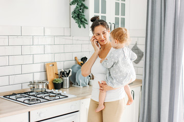 Young woman mom with baby girl on hands using mobile on bright kitchen at home