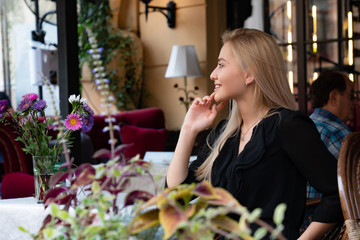 Side view portrait of a luxurious elegant blonde woman in black dress sitting at a table in a cozy cafe decorated with flowers