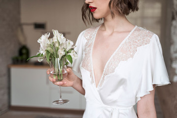 Charming modest lady in white dress with lace looking away and dreaming while holding glass with small white flowers and enjoying scent