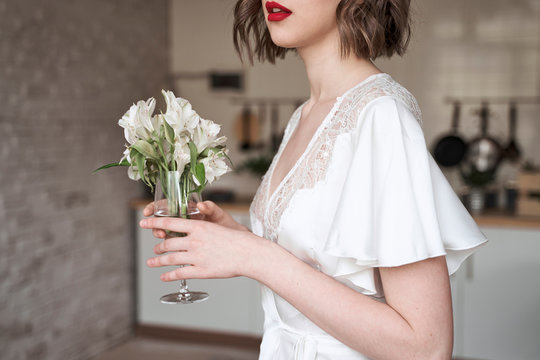 Charming modest lady in white dress with lace looking away and dreaming while holding glass with small white flowers and enjoying scent