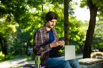 Young freelancer with beard and mustache sitting with a laptop and coffee in a park
