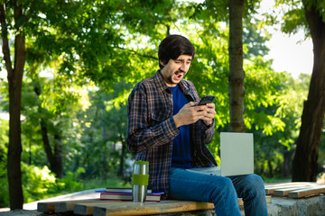 Young brunet freelancer with beard and mustache sitting with a laptop and coffee in a park.