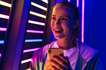 Image of woman holding smartphone by multicolored neon illumination indoors