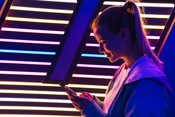 Image of woman holding smartphone by multicolored neon illumination indoors