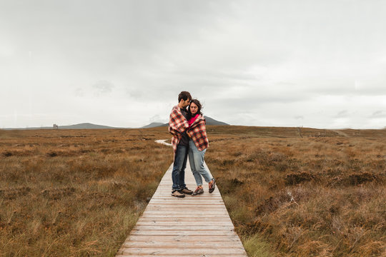 Couple of young travelers covered with plaid standing on wooden pathway among brown peat bog in Scotland on gloomy cloudy day 
