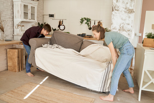 Side view of diligent man and woman barefoot carrying cozy soft coach with plaid into light kitchen
