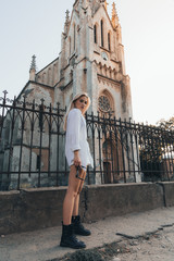 Fototapeta na wymiar Street style photo, attractive blonde girl in white shirt and black boots stands in front of the old church