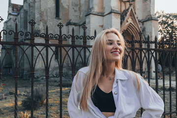 Fashionable beautiful blonde girl with loose hair, wearing white shirt, black forged fence on the...