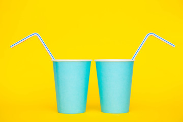 Two blue paper cups with drinking colored plastic straws on yellow background. Set for party. Minimalist Style. Copy, empty space for text
