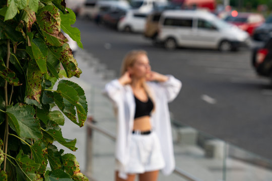 Out of focus blonde woman in white shirt and shorts standing near car parking and styling her loose hair