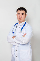 A young Asian doctor in a white coat, with a stethoscope on his neck stands holding his hands on his chest alone on a white background.