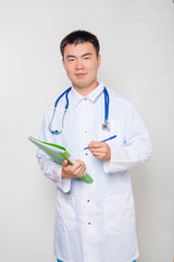 A young Asian doctor in a white coat, with a stethoscope on his neck, holds a green folder in his hands and takes notes. One on a white background.