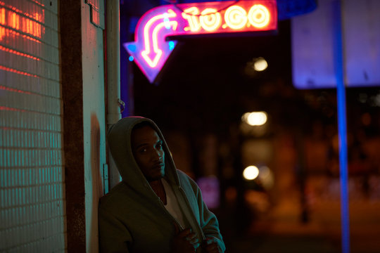 Man in hoodie standing outdoors under neon sign at night