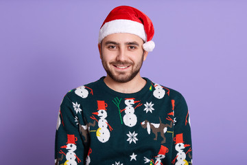 Bearded man with dark beard. Christmas holidays. Christmas decoration. Happy bearded man wearing Santa hat and sweaterwith snowman. Christmas hat. Male posing isolated over lilac background.