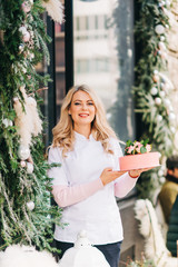 Portrait of cheerful baker woman standing next to facade of Tea Room Bakery, holding delicious pink cake with fresh flowers