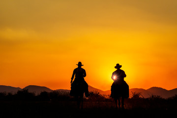 Fototapeta na wymiar Silhouette image of two cowboys riding horseback at sunset with mountain range background