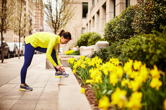A female runner tying her shoe near daffodils on a city street.