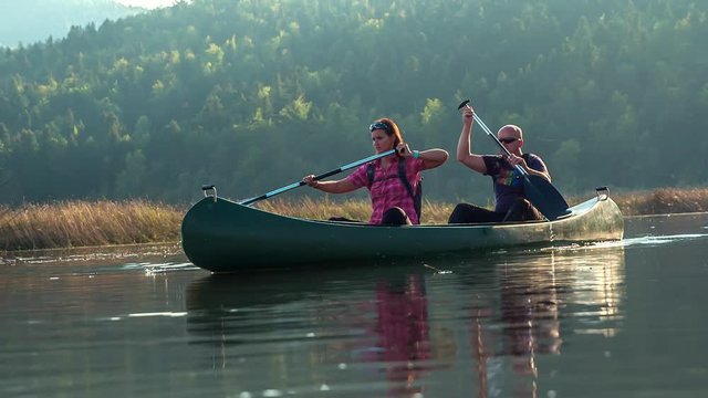 A beautiful scene of two people riding a canoe. The woman is paddling in the front of a canoe and a man is paddling in the back of a canoe. Water is peaceful and nature is outstandingly gorgeous.