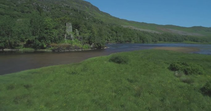 Low aerial flying over grassy sunlit marshes towards the ancient tower house ruins of Loch Dochart Castle in the Scottish Highlands