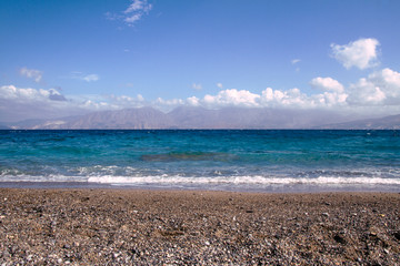 Pebble beach, turquoise water of Mediterranean Sea with white wave, mountains in the distance, beautiful sky with clouds. Seascape background. Agios Nikolaos, Crete Island. Greece