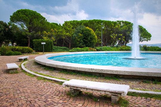 Pine trees, fountain and stone benches located on city Barcola beach in Trieste, Italy. Summer day.