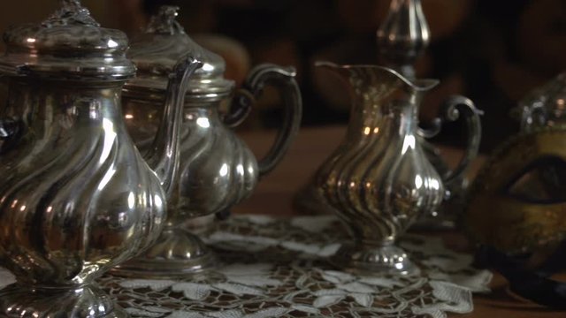 A close up slow panning on a vintage silverware teapot, milk pan and sugar bowl near ancient decorative venetian carnival mask FDV