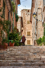 Fototapeta na wymiar Elderly woman climbing stone stairs in Mallorcan village. Street with ornamental plants. Fornalutx, Mallorca