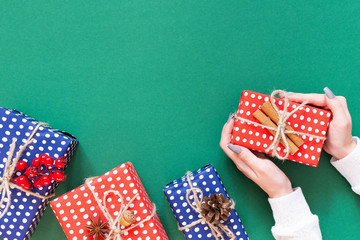 Girl holds gift box, red and blue gift boxes in polka dots with christmas tree cone and twigs of hawthorn with acorn and cinnamon on green background, flat lay, top view © pundapanda