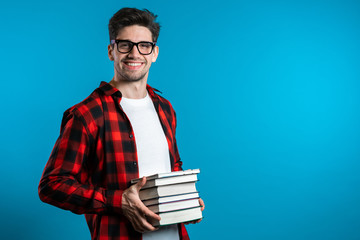Fototapeta European student in red plaid shirt on blue background in studio holds stack of university books from library. Guy smiles, he is happy to graduate. obraz