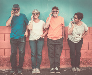 Cheerful group of four senior people in friendship standing against an orange and blue wall smiling using cell phone. Happiness and complicity