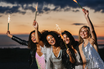 Four happy women with sparklers at sunset. Smiling stylish friends having fun outdoors.