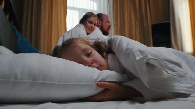 A family in the hotel room - a little girl sleeping in bed - her mom and dad looking at her