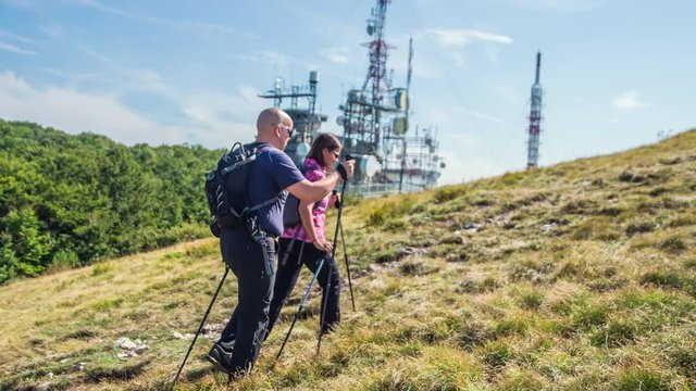 A couple is looking around when hiking and they're observing other mountains. The weather is really nice. There is also a television trasnmitter in the background.