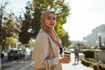 Muslim woman on a city street holds coffee in her hand and looks with an open look at the camera