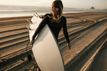 Naklejka premium Uśmiechnięta kobieta z deską surfingową spaceru na plaży podczas zachodu słońca