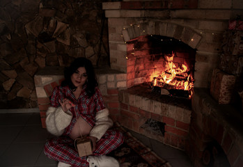 Pregnant woman dressed pajamas and warm knitted cardigan relax in room with brick fireplace with burning fire against the background of x-mas presents and lights.  Woman holding the present