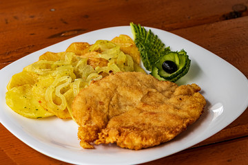 Home-style fried potatoes and a large piece of meat on a white plate. Curly chopped green cucumber. On a wooden table.