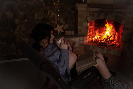 Back view of pregnant woman wearing knitted warm sweater sitting in rocking chair. Female reading the book against the burn fire in brick fireplace at home and holding a cup of tea