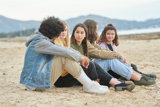 Five stylish teens sitting on sand on beach of lake.
