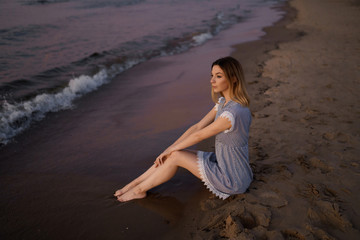 Fototapeta na wymiar Sitting: Portrait of a Beautiful blonde woman in a light blue dress on the Baltic Sea beach during sunset with vivid colors