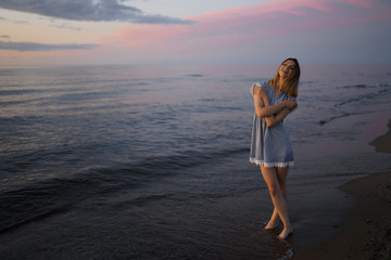 Standing full height: Portrait of a Beautiful blonde woman in a light blue dress on the Baltic Sea beach during sunset with vivid colors