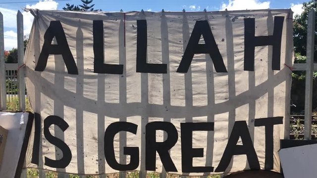 hand held shot of ragged white banner with black coloured religious text Allah is great tied on a steel fence in poor urban area on a sunny day. featuring religious statement and demonstration.