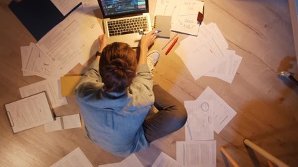 Flat lay of young Caucasian woman sitting on floor near laptop and working with papers in the evening