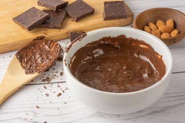 Melted chocolate in a bowl, pieces of chocolate over a wooden board, melted chocolate on a wooden spoon and almonds.