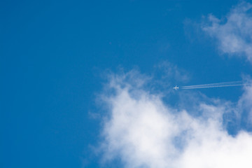 Naklejka na ściany i meble A distant airplane leaving a steam tail in a cloudy and blue sky framed by some clouds