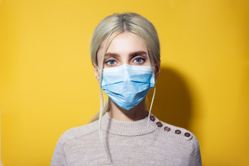 Portrait of young blonde girl with blue eyes, wearing flu medicine mask and sweater, isolated on yellow background.