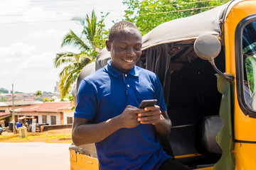 cheerful african man standing next to his tuk tuk taxi smiling and using his smart phone