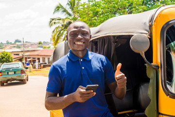 cheerful african man standing next to his tuk tuk taxi smiling and using his smart phone giving a...