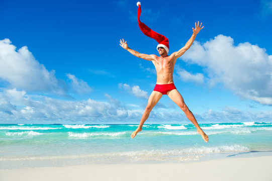 Fit Santa with extra long hat doing a Christmas celebration jump in red speedos on the shore of a tropical beach