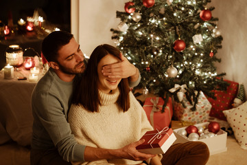 man close womans eyes and giving surprise gift her; indoor photo with xmas decoration christmas tree on background